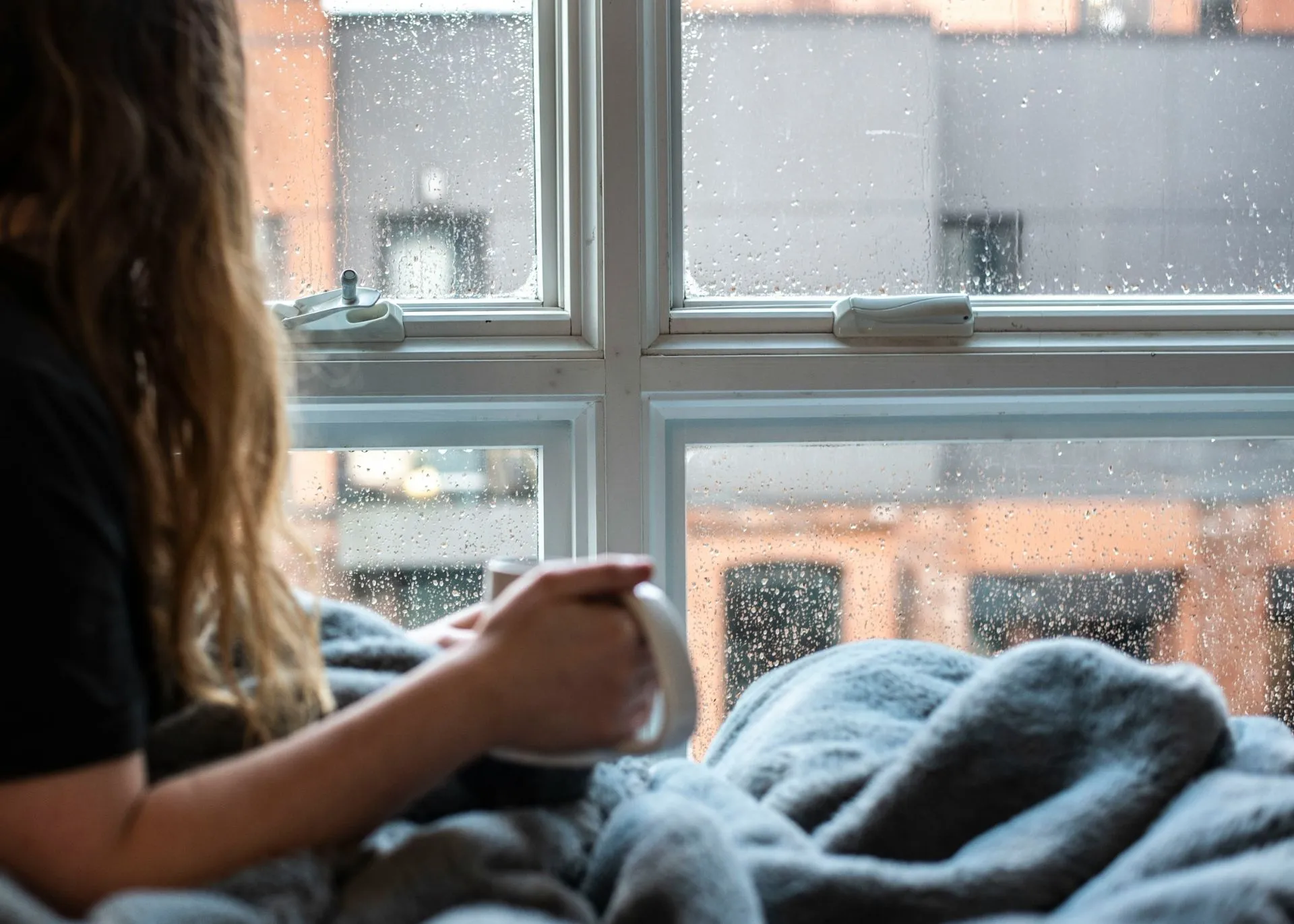 Frau sitzt mit Decke und Tasse am Fenster und schaut hinaus