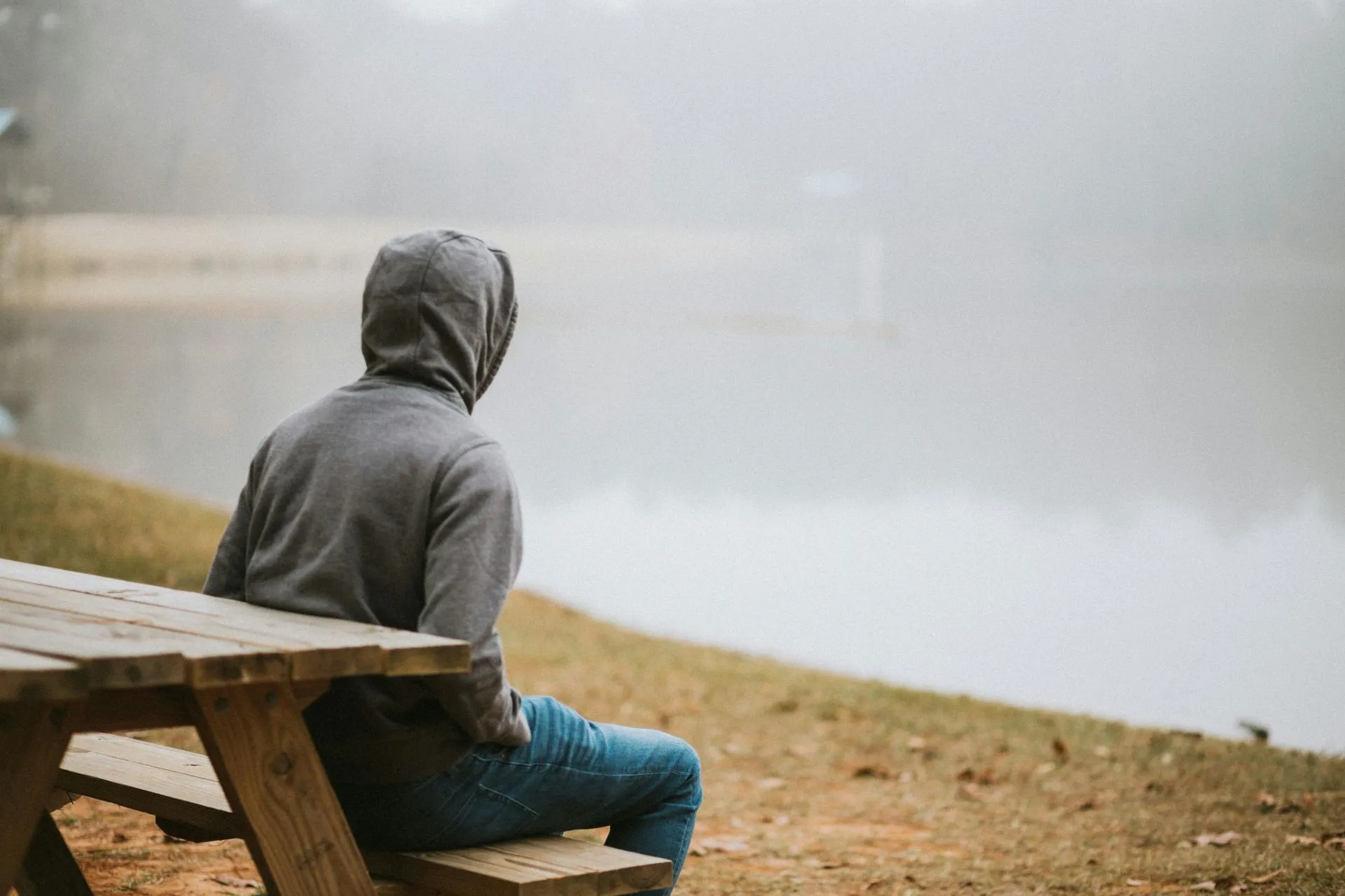 Mann im grauen Kapuzenpullover sitzt auf einer Bank und blickt auf einen Fluss im Nebel